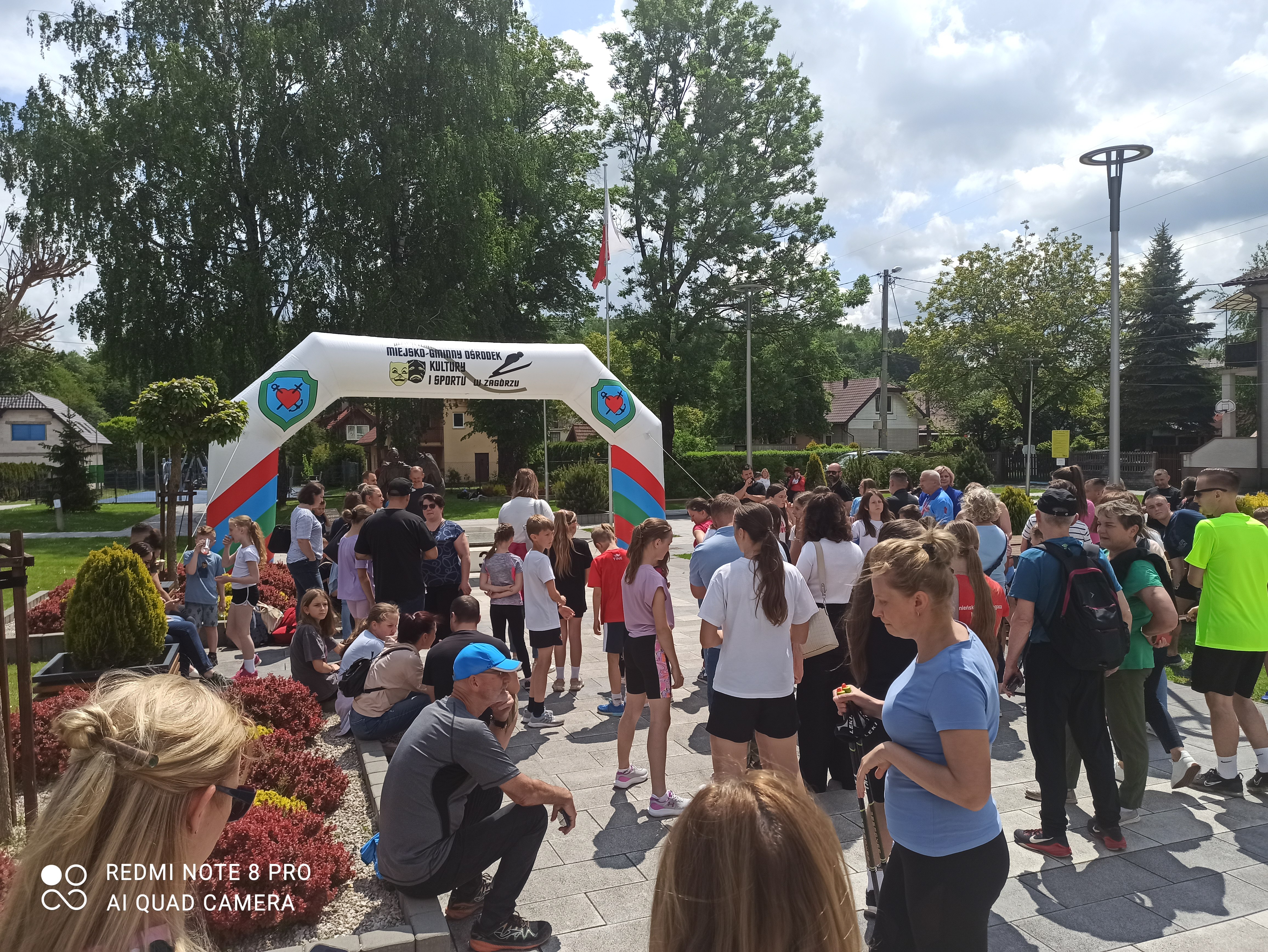 Tłum ludzi zbiera się pod łukiem z napisem "Miejsko-Gminny Ośrodek Kultury i Sportu". Wydarzenie na świeżym powietrzu, zawody